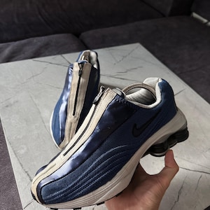 May include: A pair of blue and beige athletic shoes with a zipper closure. The shoes feature a mesh design and a black Nike logo. The soles are white and black. The shoes are approximately 11 inches long.