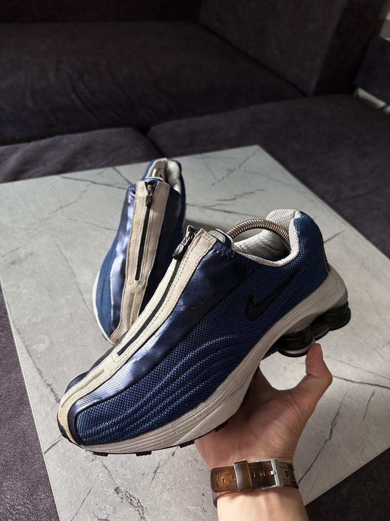 May include: A pair of blue and beige athletic shoes with a zipper closure. The shoes feature a mesh design and a black Nike logo. The soles are white and black. The shoes are approximately 11 inches long.