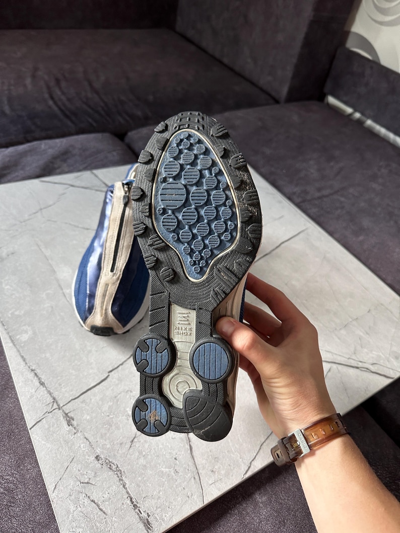 May include: A pair of blue and beige athletic shoes with a black and blue sole. The sole features a textured design with circular and oval patterns. The shoe has a zipper on the side and the brand name "Nike Shox" is visible.