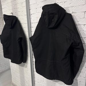 May include: A solid black hooded jacket featuring long sleeves, a structured hood, and a horizontal seam detail across the back. The garment is displayed from a rear perspective, showing a clean, uniform matte finish and a simple, functional design.