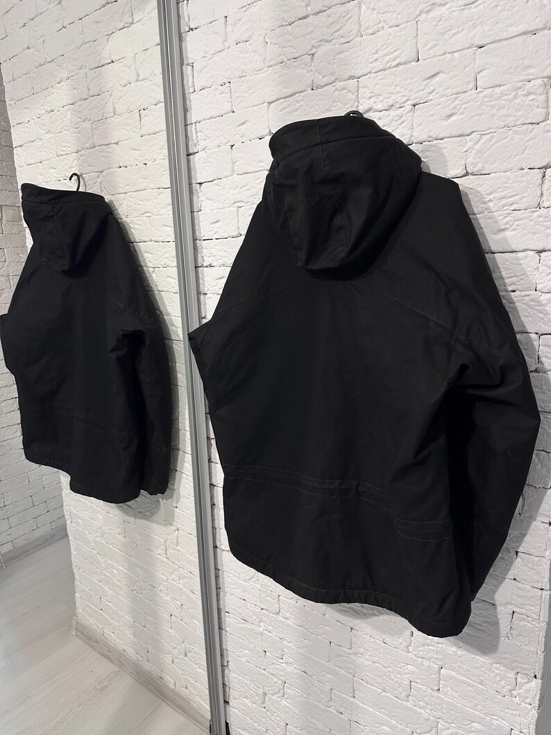 May include: A solid black hooded jacket featuring long sleeves, a structured hood, and a horizontal seam detail across the back. The garment is displayed from a rear perspective, showing a clean, uniform matte finish and a simple, functional design.