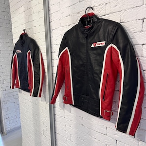May include: Black and red motorcycle jacket with white accents. The jacket features a stand-up collar, a zipper closure, and a logo on the chest. The sleeves and sides have red panels with white stripes. The jacket is hanging on a black hanger.