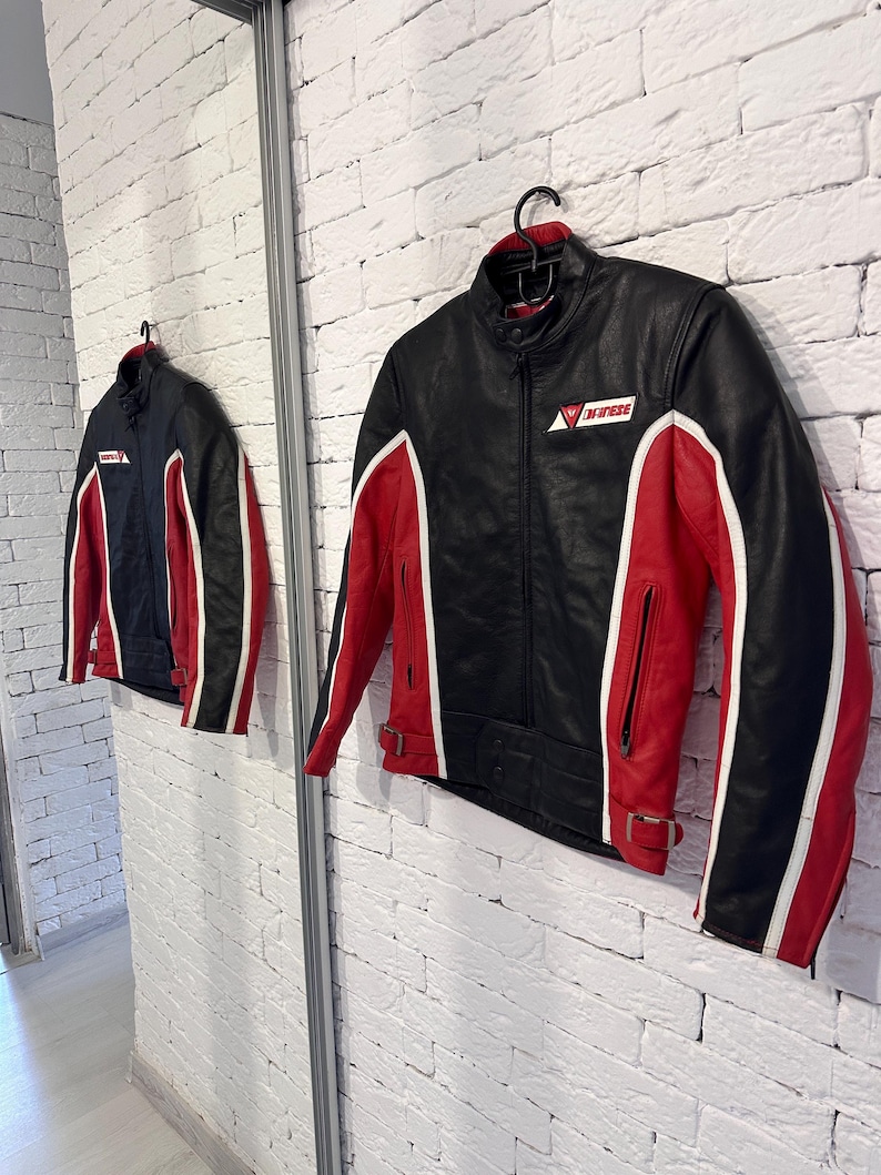 May include: Black and red motorcycle jacket with white accents. The jacket features a stand-up collar, a zipper closure, and a logo on the chest. The sleeves and sides have red panels with white stripes. The jacket is hanging on a black hanger.