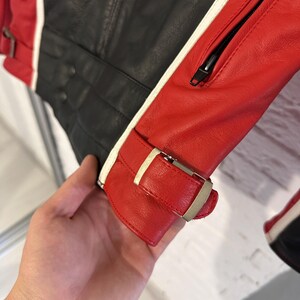 May include: A close-up of a black and red leather jacket with white stripes. The jacket has a zipper and a buckle closure on the sleeve. The leather appears to be textured.