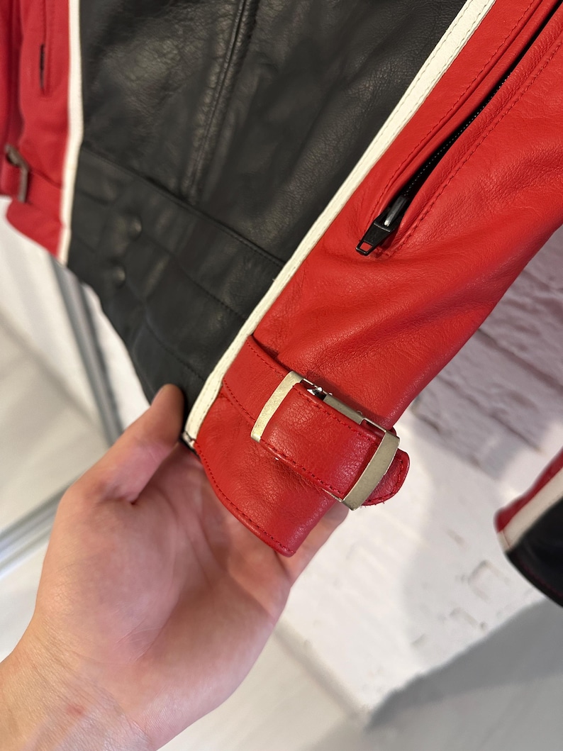 May include: A close-up of a black and red leather jacket with white stripes. The jacket has a zipper and a buckle closure on the sleeve. The leather appears to be textured.