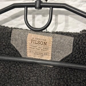 May include: A dark charcoal gray fleece-lined garment hanging on a black plastic hanger. A tan rectangular fabric label is stitched to a gray felt patch, featuring the text A GENUINE FILSON, MIGHT AS WELL HAVE THE BEST, and C.C. FILSON CO. SEATTLE.