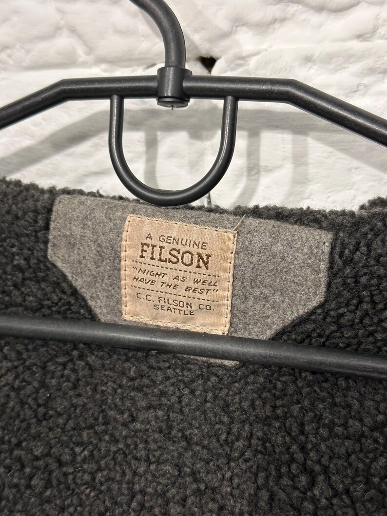 May include: A dark charcoal gray fleece-lined garment hanging on a black plastic hanger. A tan rectangular fabric label is stitched to a gray felt patch, featuring the text A GENUINE FILSON, MIGHT AS WELL HAVE THE BEST, and C.C. FILSON CO. SEATTLE.