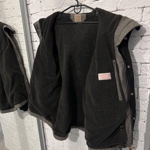 May include: A black and grey hooded jacket featuring a dark charcoal grey fleece lining. The jacket includes a front zipper closure with metal snap buttons, a hood, and a rectangular brand label sewn into the interior neck and chest area.