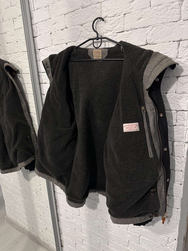 May include: A black and grey hooded jacket featuring a dark charcoal grey fleece lining. The jacket includes a front zipper closure with metal snap buttons, a hood, and a rectangular brand label sewn into the interior neck and chest area.