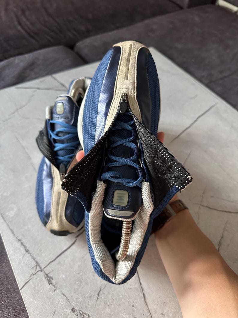 May include: Pair of blue and beige athletic shoes with a zipper closure. The shoes have blue laces and a beige suede-like material on the upper. The zipper reveals the laces and tongue. The shoes are on a marble surface.