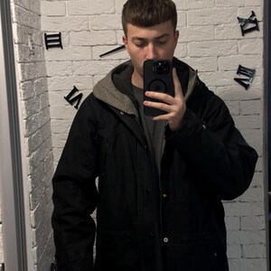 May include: A black hooded jacket featuring a grey inner lining, a front zipper closure, and metallic snap buttons. The jacket is worn over a dark grey t-shirt and light blue denim jeans, with a black smartphone held in front of the chest.
