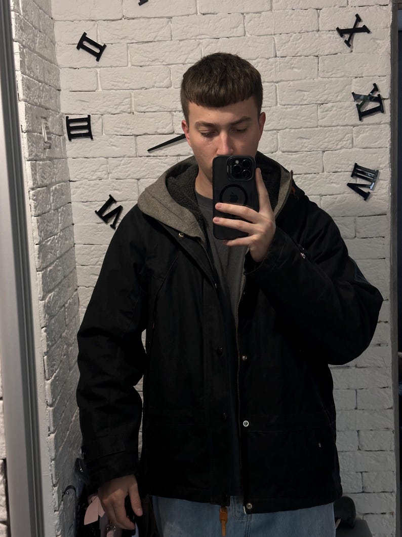 May include: A black hooded jacket featuring a grey inner lining, a front zipper closure, and metallic snap buttons. The jacket is worn over a dark grey t-shirt and light blue denim jeans, with a black smartphone held in front of the chest.