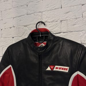 May include: Black leather motorcycle jacket with red and white accents. Features a stand-up collar, a zipper closure, and the Dainese logo on the chest. The jacket is hanging on a black hanger against a white brick wall.