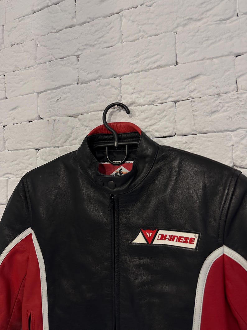 May include: Black leather motorcycle jacket with red and white accents. Features a stand-up collar, a zipper closure, and the Dainese logo on the chest. The jacket is hanging on a black hanger against a white brick wall.