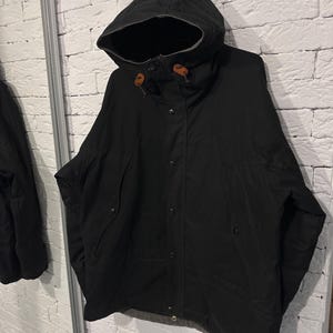 May include: A black hooded jacket featuring a front button closure, a high collar, and two slanted side pockets. The hood includes brown leather toggles at the drawstring ends, and the jacket has a visible grey lining along the bottom hem.