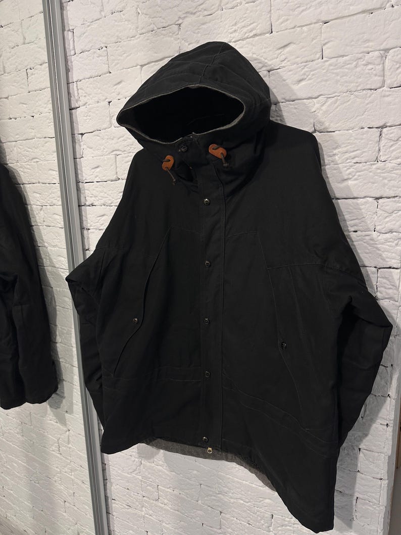May include: A black hooded jacket featuring a front button closure, a high collar, and two slanted side pockets. The hood includes brown leather toggles at the drawstring ends, and the jacket has a visible grey lining along the bottom hem.