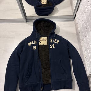 May include: A navy blue zip-up hoodie with a dark brown faux fur lining. The front features the white text "HOLLISTER 19" on the left and "STER 22" on the right. A Hollister California brand label is sewn inside the neck. It has long sleeves and a hood.