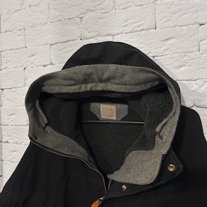May include: A black hooded jacket featuring a grey fleece-lined interior and a hood. The garment includes a tan leather zipper pull and a rectangular label on the inner collar with the text FILSON printed in black capital letters.