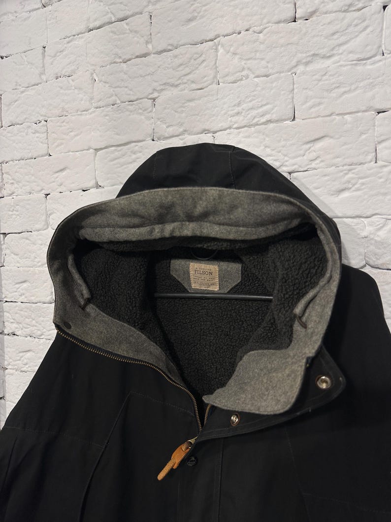 May include: A black hooded jacket featuring a grey fleece-lined interior and a hood. The garment includes a tan leather zipper pull and a rectangular label on the inner collar with the text FILSON printed in black capital letters.