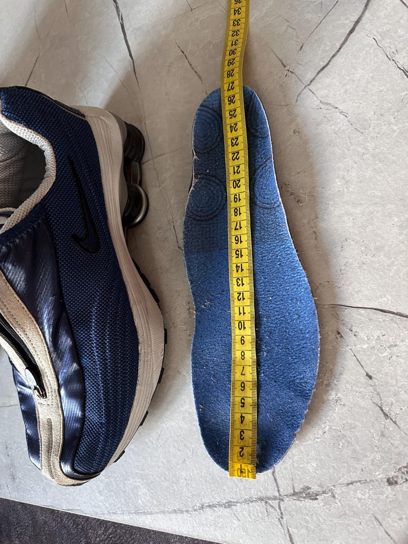 May include: A blue and white athletic shoe next to a blue insole being measured with a yellow tape measure. The insole measures approximately 10.5 inches in length.