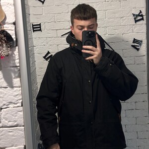 May include: A black hooded jacket featuring a front zipper closure, a snap-button placket, and brown leather-like toggle details at the collar. The jacket has long sleeves with adjustable cuffs and a structured hood, providing a solid black finish.