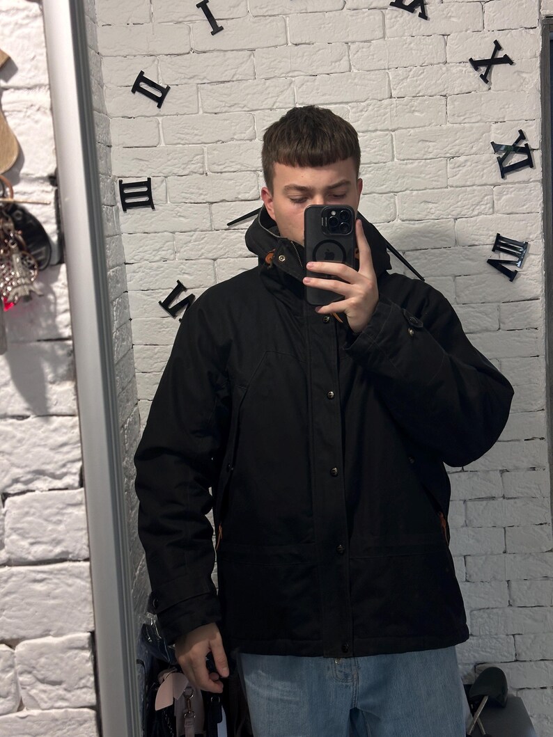 May include: A black hooded jacket featuring a front zipper closure, a snap-button placket, and brown leather-like toggle details at the collar. The jacket has long sleeves with adjustable cuffs and a structured hood, providing a solid black finish.