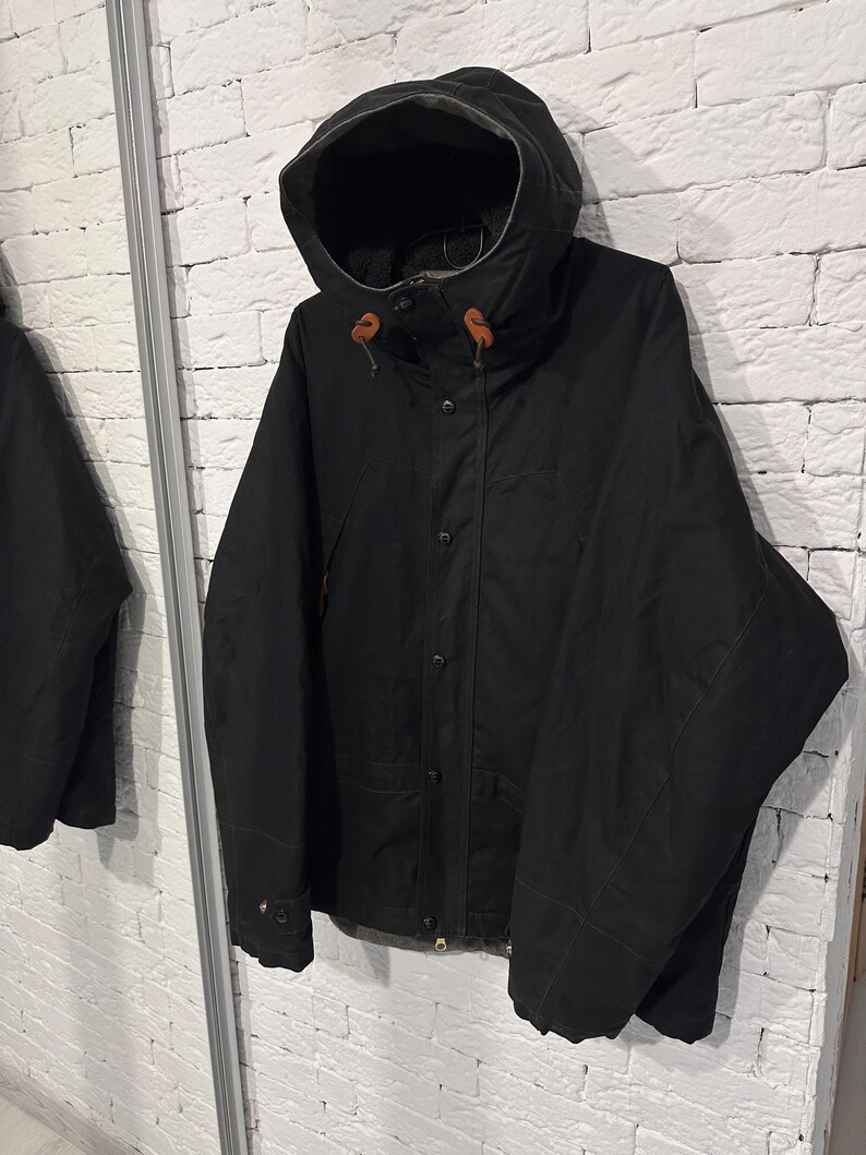 May include: A black hooded jacket featuring a front button closure, long sleeves, and brown leather toggles on the hood drawstrings. The jacket has a structured collar, a visible interior lining, and a clean, solid-colored exterior design.