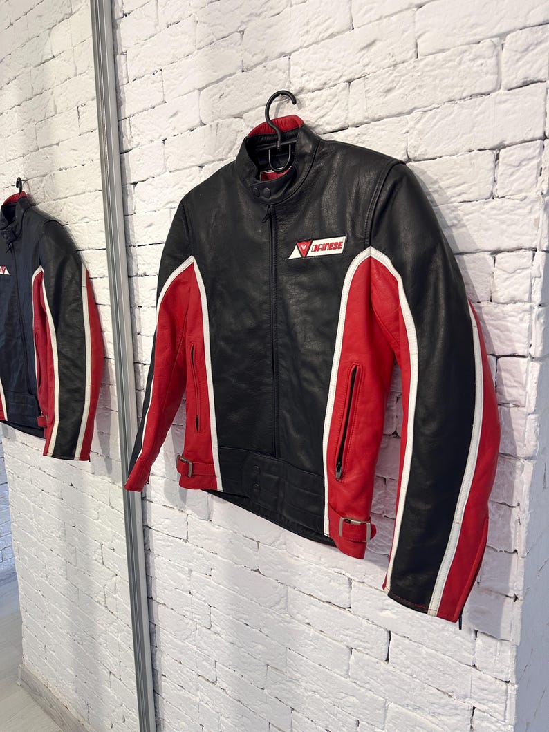 May include: Black and red leather motorcycle jacket with white accents. The jacket features a stand-up collar, zippered pockets, and a logo on the chest. The sleeves and sides are red with white stripes.
