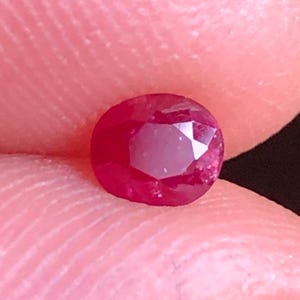 Può includere: Una piccola gemma di rubino sfaccettata e rotonda. La gemma è di un colore rosso intenso e ricco, con strutture interne visibili. Il rubino è tenuto tra due dita, mostrando le sue dimensioni e la sua brillantezza. Lo sfondo è sfocato.
