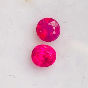 May include: Two round, faceted, bright pink gemstones. The gems are set against a white background. The stones have a high luster and appear to be of high quality. The gems are likely rubies or pink sapphires.