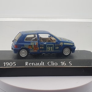 Puede incluir: Un coche modelo de fundición a presión Renault Clio 16 S azul de 1905 en una vitrina transparente. El coche tiene un número 91 blanco y la palabra "elf" en el lateral.