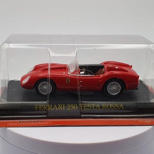 Op de afbeelding: Een rode Ferrari 250 Testa Rossa model auto in een transparante kunststof display case. De auto staat op een zwarte basis met de woorden "Ferrari 250 Testa Rossa" in goud gedrukt.