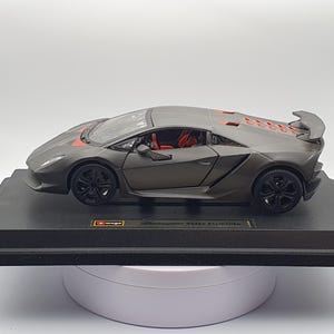 Może przedstawiać: Szary model samochodu Lamborghini Sesto Elemento z czerwonymi akcentami, czarnymi kołami i tylnym spojlerem. Model stoi na czarnym stojaku ekspozycyjnym. Wnętrze samochodu jest czerwone.