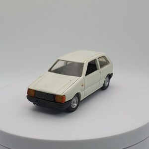 May include: A die-cast model of a white hatchback car. The toy car has black tires, a black front grill, and orange-tinted headlights. The car is sitting on a white, circular surface.