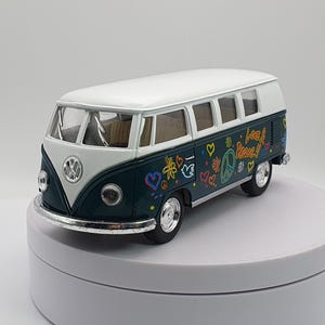 Può includere: Un autobus Volkswagen in miniatura verde acqua e bianco con le parole "Love & Peace" e scarabocchi colorati. L'autobus giocattolo ha un tetto bianco, finiture cromate e un logo VW sul davanti.