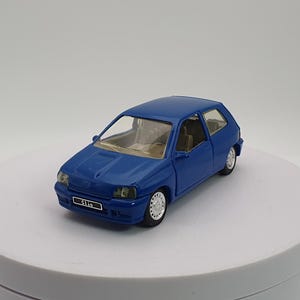 Può includere: Un'auto giocattolo blu di piccole dimensioni con un design hatchback. L'auto modello ha interni dettagliati e ruote bianche. L'auto è su una superficie bianca. La targa dell'auto recita "Clio".