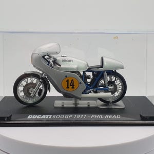 Może przedstawiać: Model motocykla Ducati 500GP w kolorze srebrnym i niebieskim w przezroczystej gablocie. Motocykl ma numer "14" na boku i napis "DUCATI 500GP 1971 - PHIL READ" na podstawie.