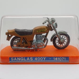 Puede incluir: Modelo en miniatura de una motocicleta Sanglas 400Y en una vitrina de plástico transparente. La motocicleta es principalmente marrón con detalles plateados y un asiento negro. El modelo se encuentra sobre una base naranja con una etiqueta azul y blanca que dice "SANGLAS 400Y - 141071".