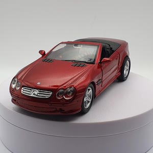 Può includere: Un modello di auto giocattolo rosso convertibile con tetto nero. L'auto ha una griglia anteriore e fari anteriori dettagliati.