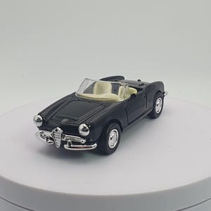 Può includere: Un'auto giocattolo cabriolet nera con interni color crema. L'auto classica ha dettagli argentati sulla parte anteriore e sulle ruote. L'auto è esposta su una superficie bianca.