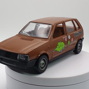 May include: A brown toy car with black trim and wheels. The car has a green cartoon graphic and the text "Uno 55S" on the side. The car is on a white surface.