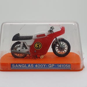 Puede incluir: Modelo en miniatura de una motocicleta Sanglas 400Y roja, blanca y negra, presentado en una vitrina de plástico transparente. El modelo tiene el número 3 en el lateral y descansa sobre una base naranja con texto.