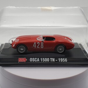 May include: A red 1:43 scale model car of an OSCA 1500 TN from 1956. The car has a white number "428" on the side. The model is displayed in a clear plastic case on a black base with the words "1000 Miglia" and "OSCA 1500 TN - 1956".