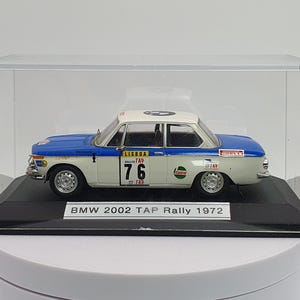 Può includere: Un'auto modello BMW 2002 bianca e blu del 1972 in una teca trasparente. L'auto ha il numero "76" rosso e bianco sul lato. La teca ha una base nera con il testo "BMW 2002 TAP Rally 1972".
