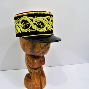 Puede incluir: Un sombrero negro y rojo con un diseño bordado en oro. El sombrero tiene una visera negra y está sentado en un expositor de madera.