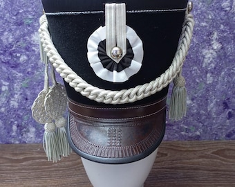 May include: A black and white military style hat with a silver cord and tassel. The hat has a black band with a white circle and a black center.