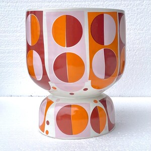 May include: A ceramic planter with a geometric design. The planter features a white base and a body adorned with alternating squares and circles in shades of orange, red, and pink. The design is symmetrical and modern.