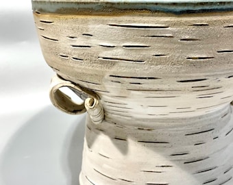 May include: A ceramic bowl with a white, textured exterior featuring horizontal black markings that mimic birch bark. The bowl has a wide rim with a blue-toned glaze, two small side handles, and contains a yellow citrus fruit.