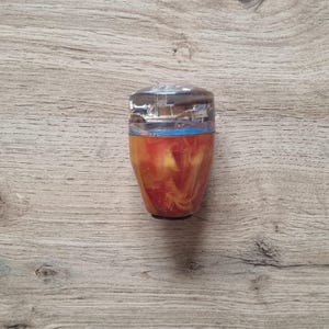 Op de afbeelding: Een decoratieve flesstopper met een doorzichtige bovenkant en een kleurrijk, wervelend interieur van rode, oranje en gele tinten. De stopper heeft een blauwe band aan de bovenkant, tegen een houten achtergrond.