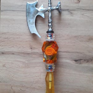 May include: A silver axe-shaped bottle opener with a clear and orange glass handle. The handle has a decorative design with a swirl pattern.